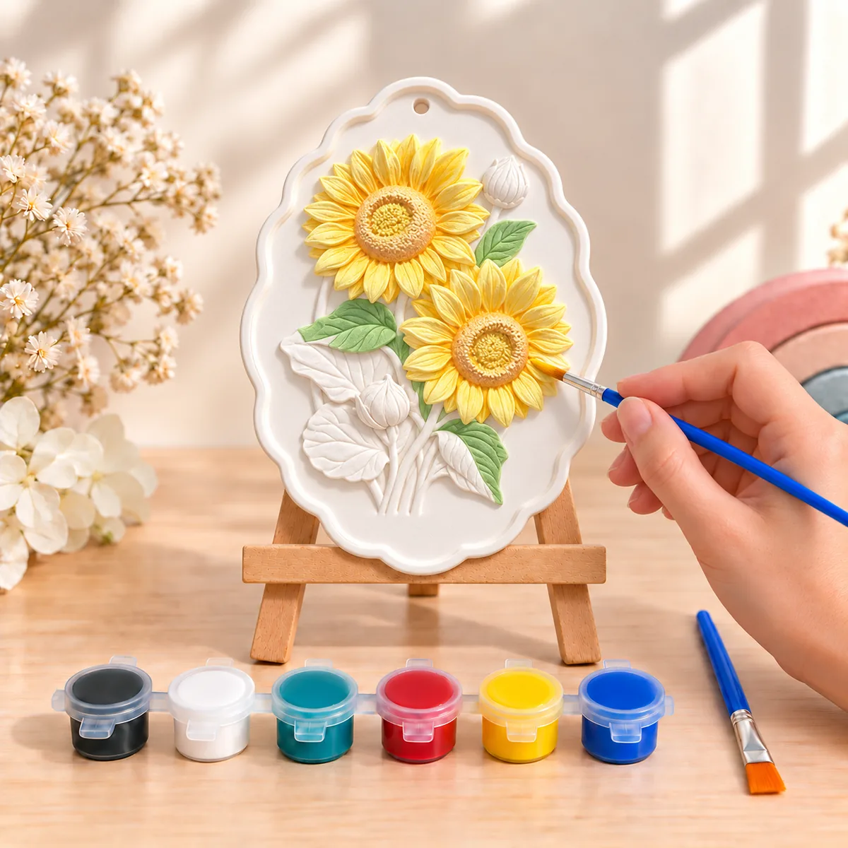 Two sunflowers in raised plaster on a mini wooden easel, petals painted yellow and leaves green, with six pots of acrylic paint and a brush in front