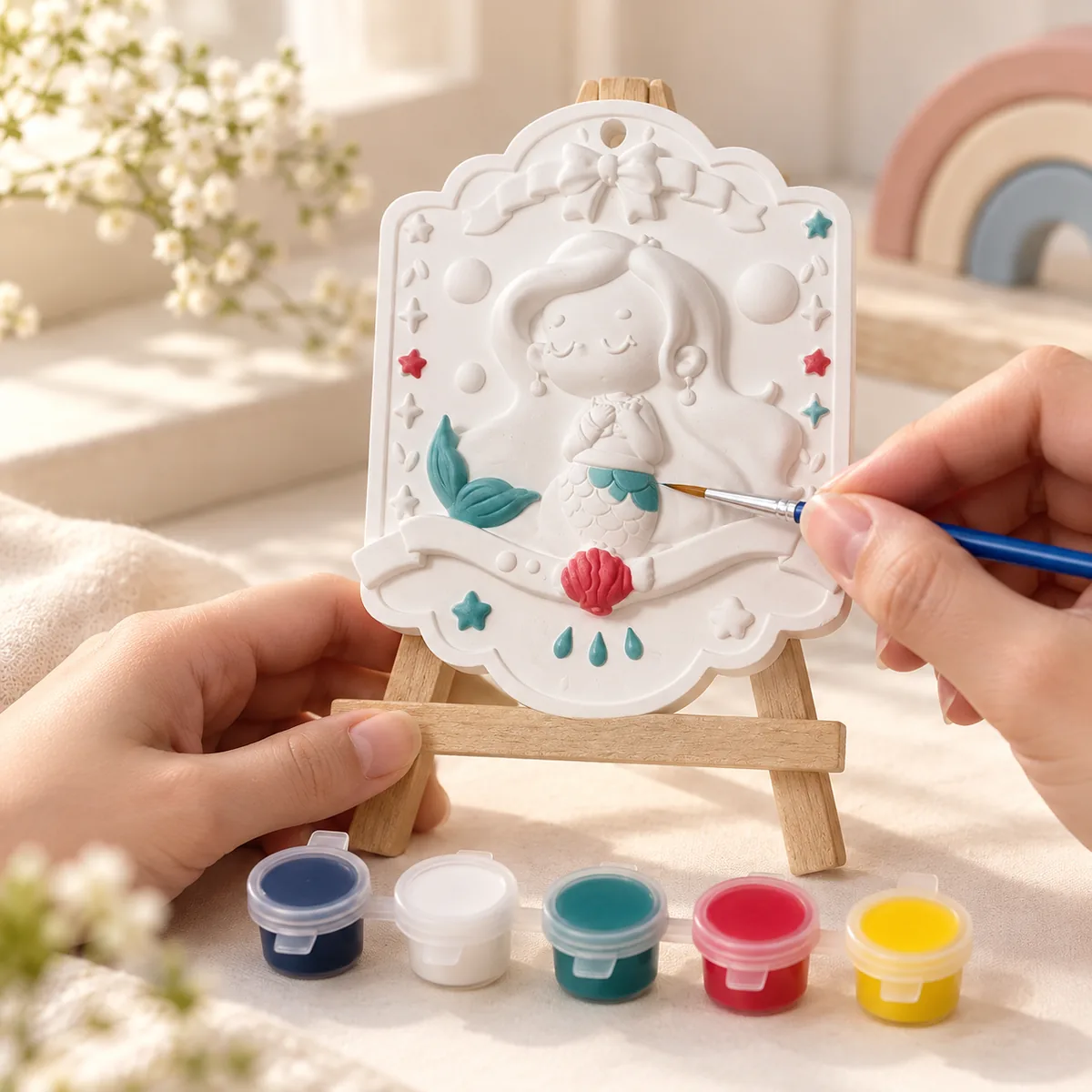 Two hands steadying a mini easel while a fine brush paints a mermaid plaque — the tail teal and the scallop shell red, the rest still bare plaster