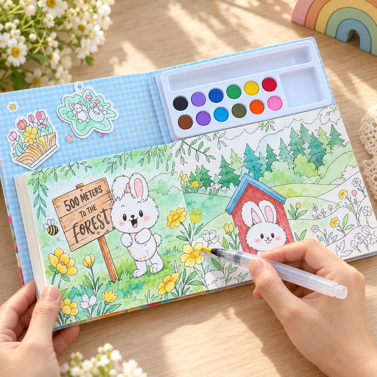 The book open flat, paint tray clipped inside the cover, a hand filling in a woodland scene with the water brush while sticker sheets sit on the left page